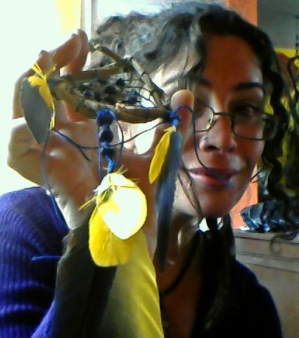 Alice wearing glasses holds up a handmade ornament with blue beads and yellow feathers, indoors.