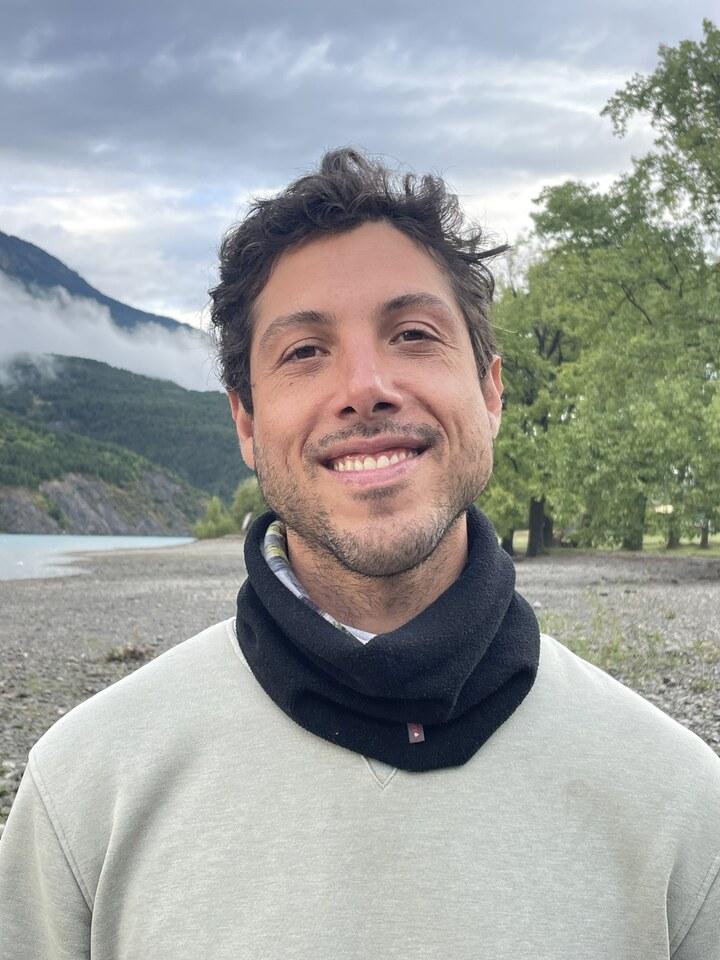 Gian smiling outdoors by a lake with misty mountains and trees in the background, wearing a light sweatshirt and black neck warmer.