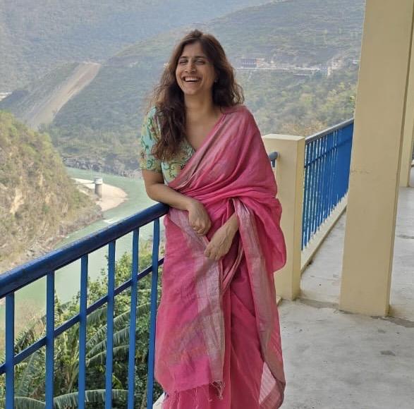 Kanicain a pink sari laughs while leaning on a blue railing on a balcony overlooking a river valley and hills.