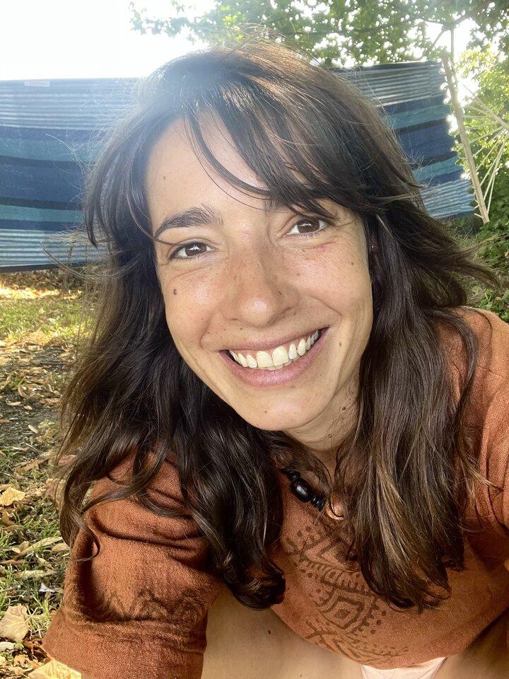 Close-up outdoor selfie of Marie with long dark hair smiling and sitting in sunlight near fallen leaves and greenery.