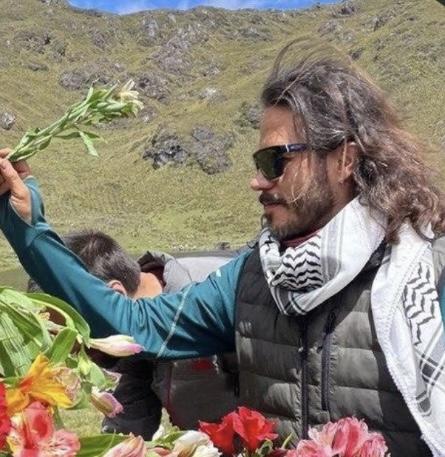 Pavel with long hair and sunglasses holds up a leafy branch near colorful flowers, with grassy mountains behind him.